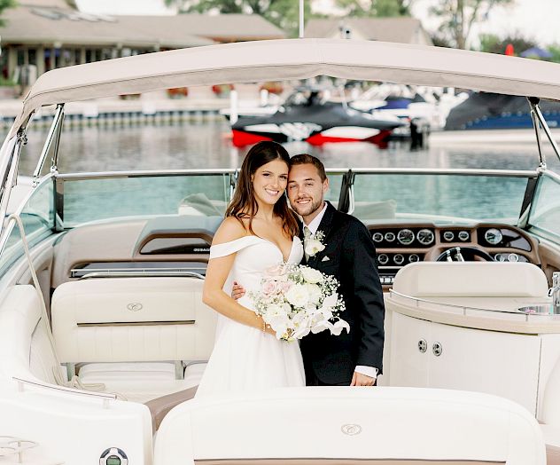 A couple in wedding attire is posing on a boat, with the bride holding a bouquet and water and docks in the background.