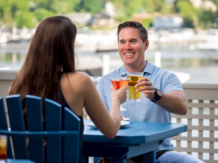 A man and a woman toast with drinks at an outdoor table near a waterfront, enjoying their time together on a sunny day, smiling happily.