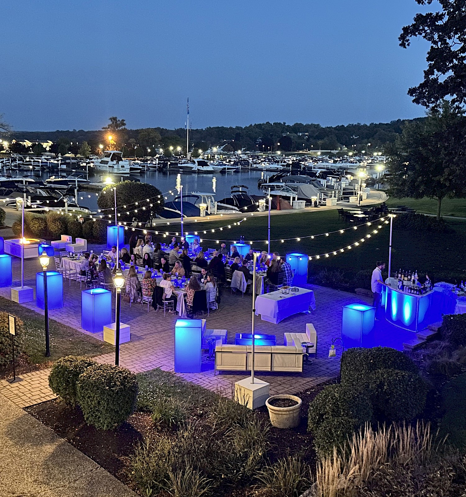 An outdoor evening event by the water, decorated with blue lights, tables, and people seated, surrounded by greenery and a marina.