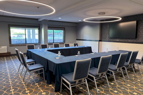 A conference room with a U-shaped table arrangement, chairs, water glasses, a window, and a wall-mounted screen.