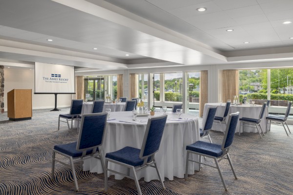 A conference room with round tables, blue chairs, a podium, and a projector screen displaying