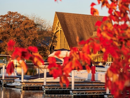 The image shows a marina with boats docked near a building with a tall, triangular roof, framed by autumn leaves with vibrant red colors.