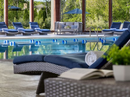 The image shows a relaxing poolside area with lounge chairs, a pool, and greenery outside. A rolled towel and open book are in the foreground.